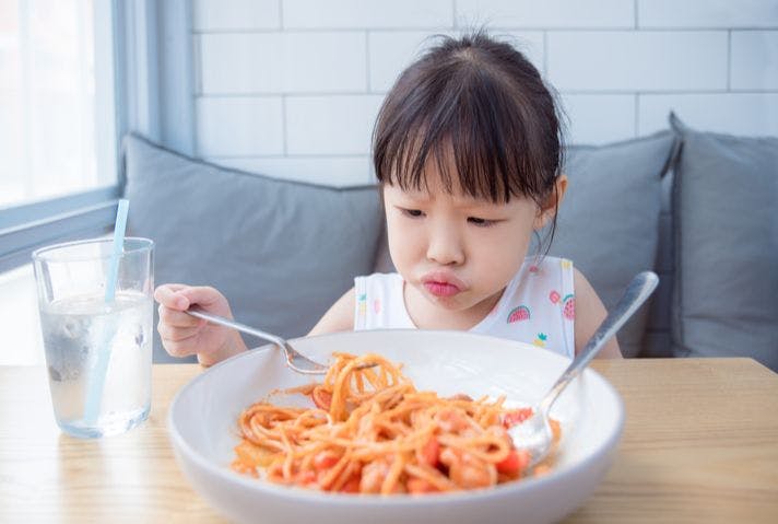 A girl with a frown on her face picking up spaghetti noodles from a bowl with a fork