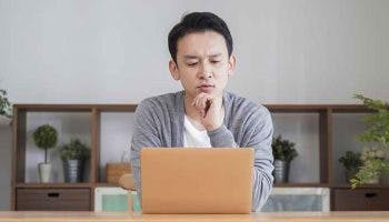 An Asian man looking confused, staring at his laptop in the living room