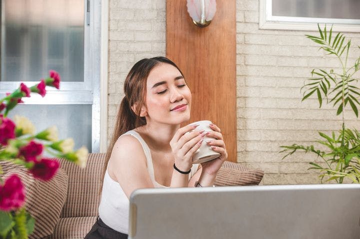 Woman smiles contently and calmly while sipping a cup of tea in front of her laptop.