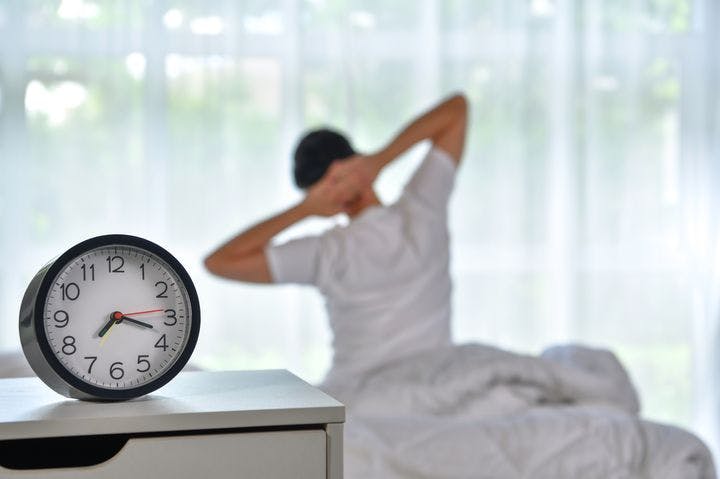 Man waking up by stretching his body facing the window, a clock shows it’s 7am