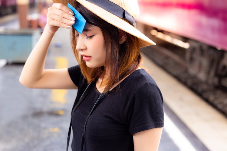 Woman with hat closing her eyes while using a blotting paper to wipe sweat off her forehead