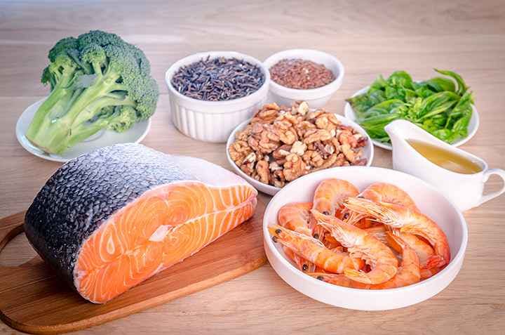 Salmon, prawns, walnuts, broccoli, basil, and grains are displayed on a wooden table.