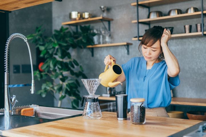 Woman looking regretful as she is about to pour coffee into a tumbler while holding her left fist to her head. 