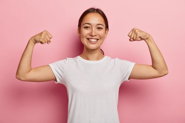 Close-up of smiling woman flexing both her arms as she stands in front of a pink wall. 