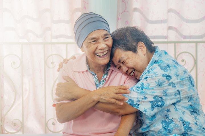 Two older women smile while hugging each other; one is a cancer patient with her head wrapped.