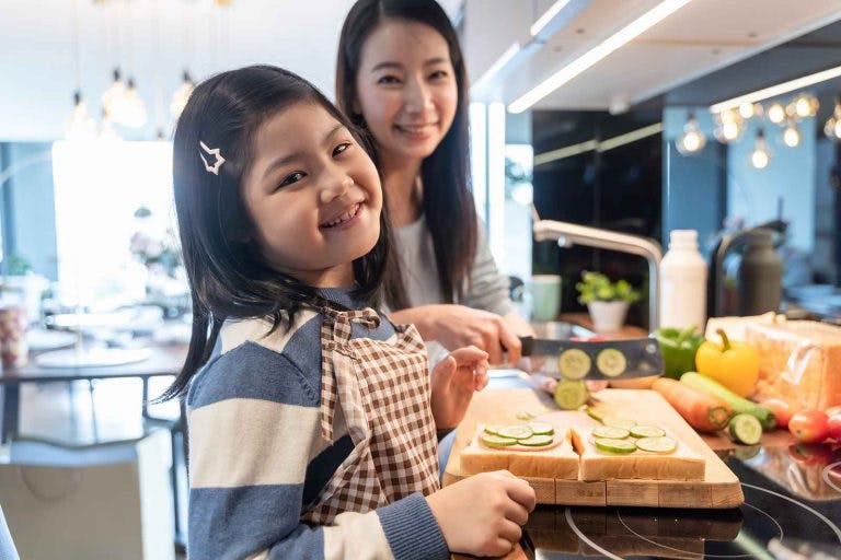 Asian mum chops vegetables while her apron-wearing young daughter helps in the kitchen.
