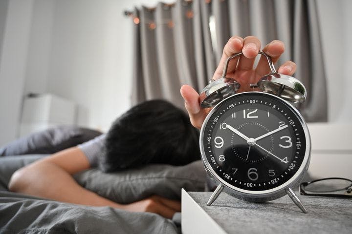 A man asleep on a bed while hugging a pillow and holding a table clock with his left hand.