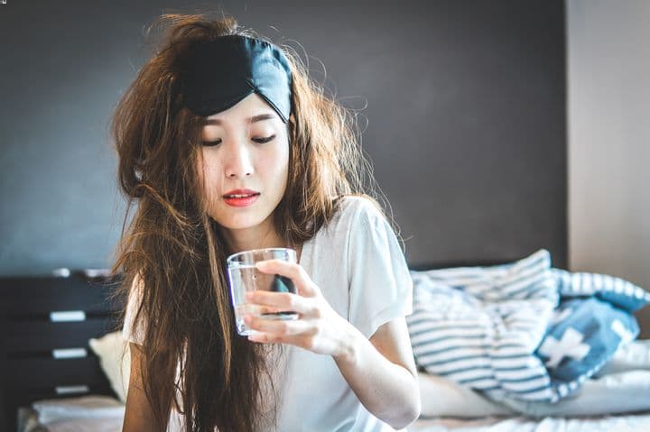 A woman sitting up in bed with a haggard appearance while holding a glass of water