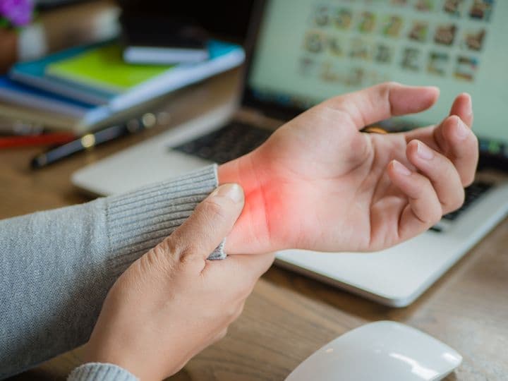 Woman holds her left wrist in pain, her laptop in the background. 