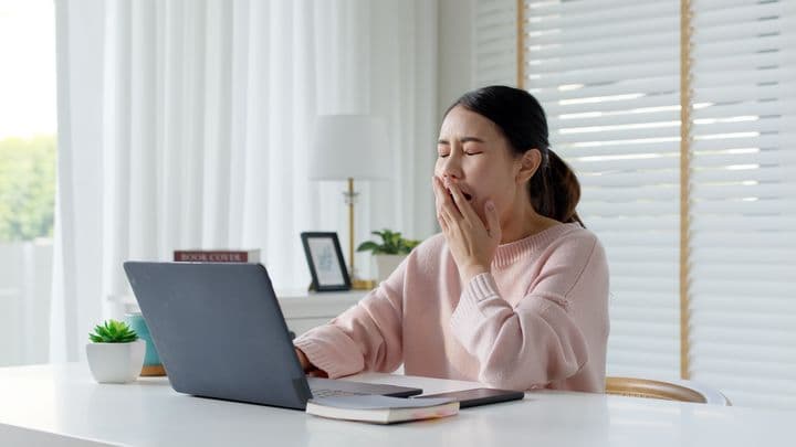 Woman yawning while covering her mouth with her left hand as she sits at a desk in front of a desktop computer.