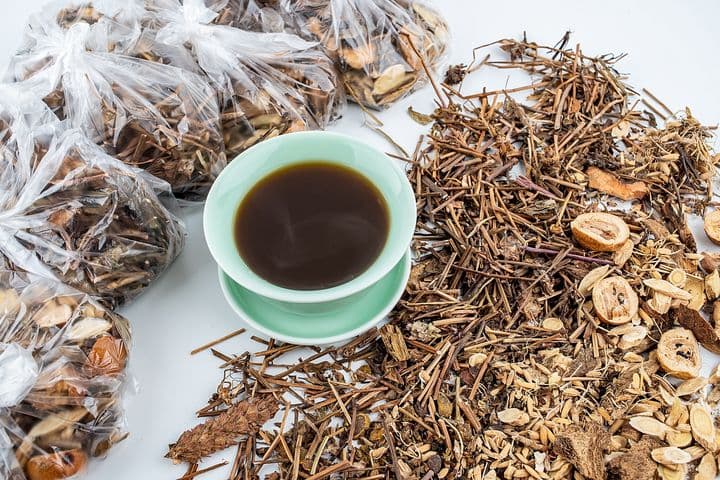 A cup containing a Chinese herbal remedy on a white surface surrounded by loose dried herbs and herbs in plastic bags.