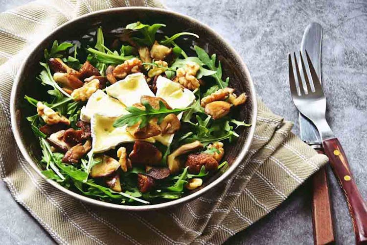 A salad plate containing brie cheese, arugula leaves, walnuts and dried figs displayed with a napkin, fork, and knife on a marble table