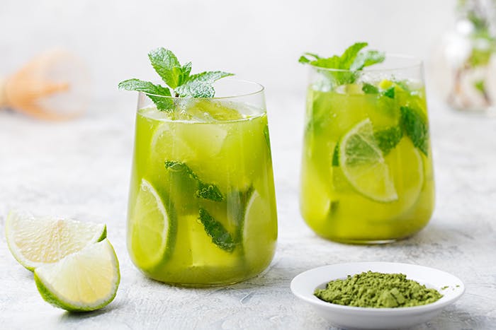 Two glasses of iced green tea with lime and mint leaves displayed alongside lime slices and matcha powder. 