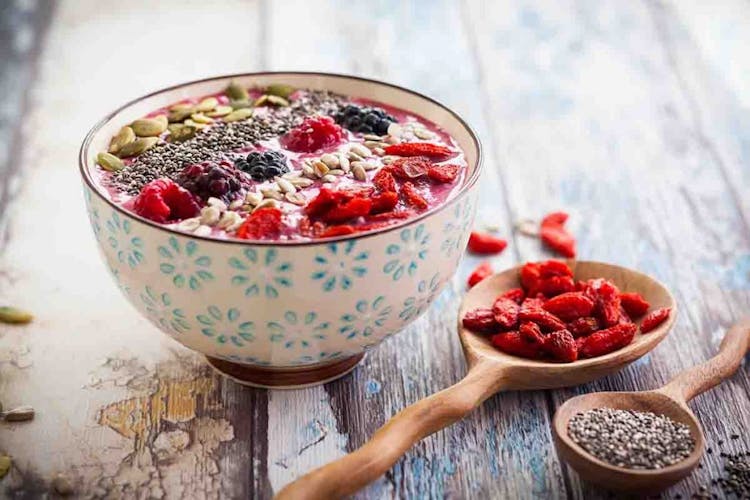 A granola bowl placed next to a spoonful of goji berries and chia seeds on a wooden table
