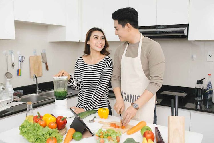 An Asian woman prepares a drink using a blender while an Asian man cuts up fruits and vegetables in a kitchen