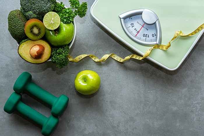 Close-up of a green weight scale, next to a pair of dumbbells, and a bowl of fruits and veggies
