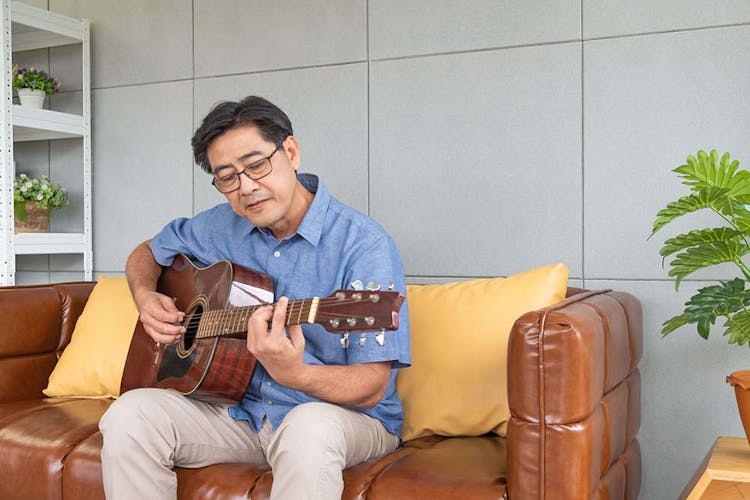 An elderly gentleman sitting on a brown leather couch playing the guitar