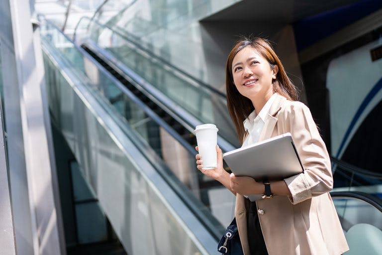 A businessperson looking up smilingly as she holds a cup of coffee in her right hand and a laptop in her left