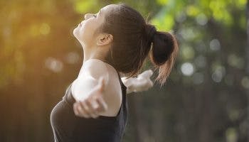 A woman in a black top raising her arms to her sides as she takes in a deep breath outdoors