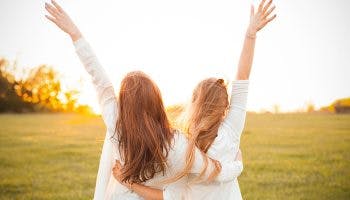 Two female best friends embracing each other’s back and lift their free hands into the air