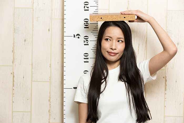 An Asian woman putting a wooden block on her head while standing in front of a height measurement scale