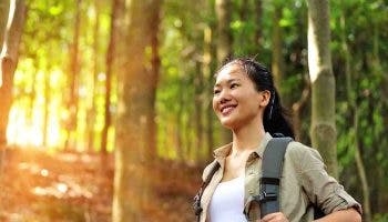 An Asian woman with a backpack hiking a mountain forest happily as the sun shines in the background