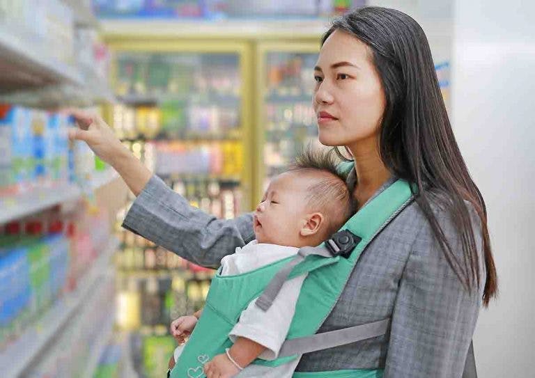 A mother carrying her baby in a front carrier while trying to choose groceries at a store