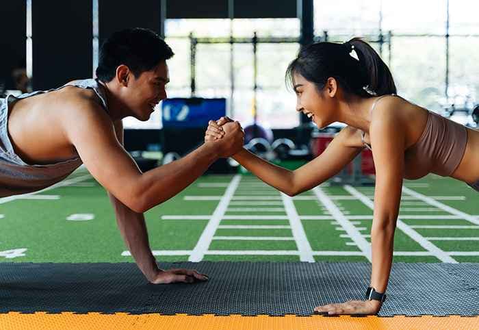 A couple in sports attire supporting each other’s weight on one arm whilst maintaining a plank position at the gym