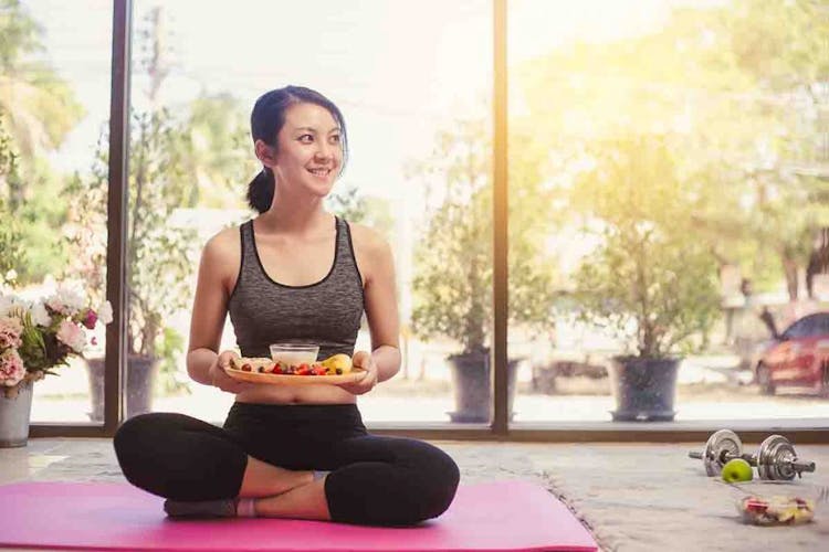 An Asian woman sitting on a yoga exercise mat while holding a plate of fruits and yoghurt