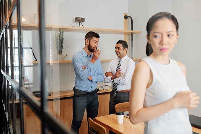 Young businesswoman standing in a meeting room listening to two businessmen talking behind her back