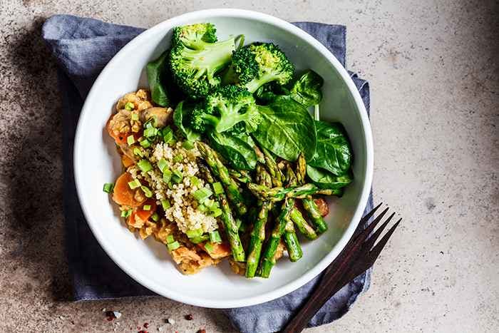 A bowl of quinoa salad mixed with other vegetables like asparagus that is placed on a blue napkin next to a fork