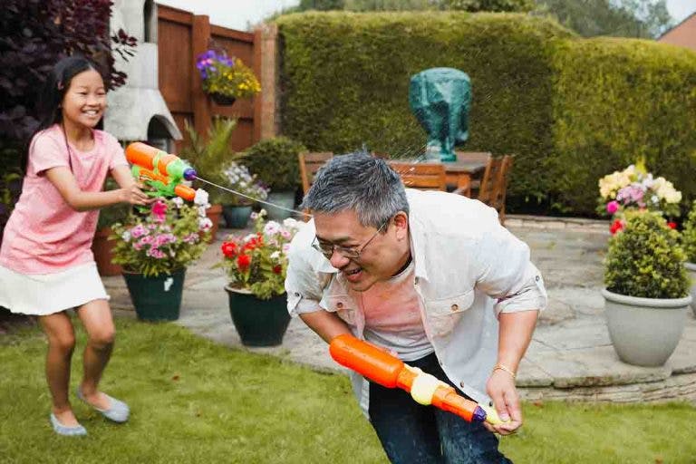 A young Asian girl shooting a water gun playfully at her father in the backyard 
