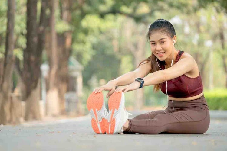 A young Asian lady sitting on the ground stretching her right hamstring, outdoor settings.