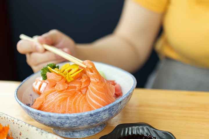 A partial view of a hand holding chopsticks and a bowl of salmon sashimi
