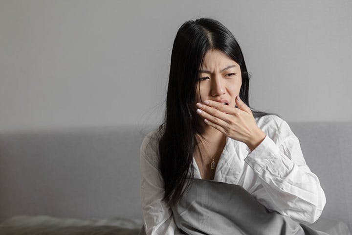 A young woman sitting up on her bed while checking her breath 