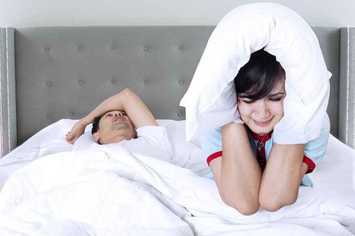 A man sleeping on a bed and snoring while his wife uses a pillow to cover her ears