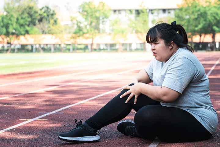 Woman with obesity sitting on a running track holding her right knee in pain.