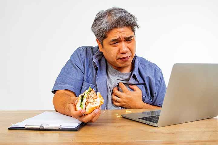 Middle-aged man eating a sandwich while working on laptop, clutching chest due to acid reflux pain