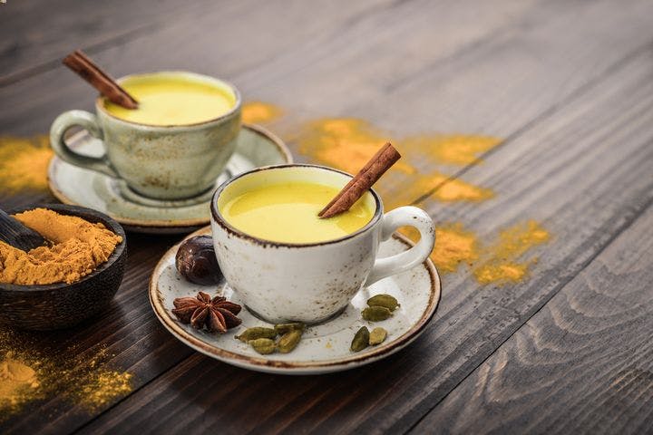 Two cups of turmeric milk on a wooden table with turmeric powder in the background