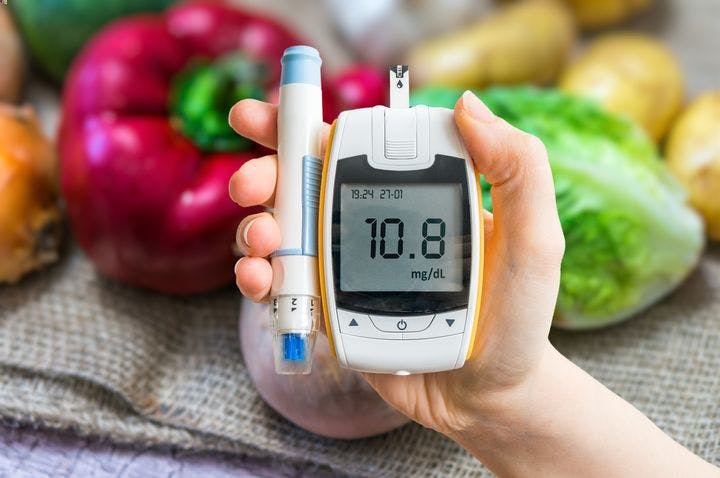 A woman holding up a blood glucose monitor with fresh vegetables in the background
