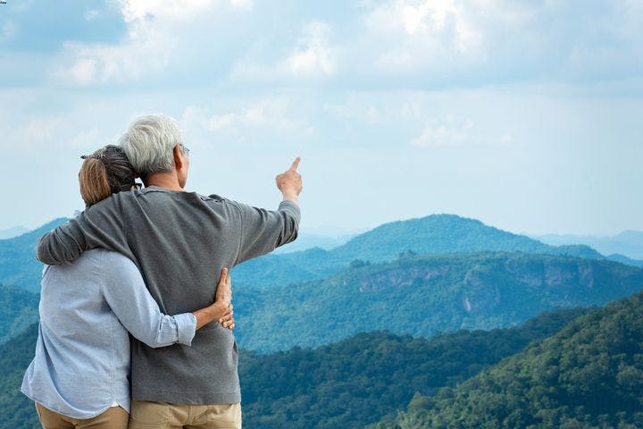 Man and woman embracing as the man points with his right finger as they look at a view of mountaintops