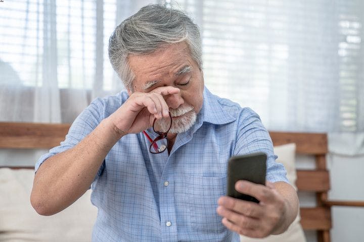 Man rubbing his eye with a finger as he holds a pair of spectacles in his right hand and mobile phone in his left