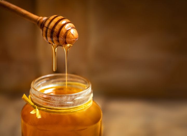 A wooden dripper being lifted from a glass jar containing honey