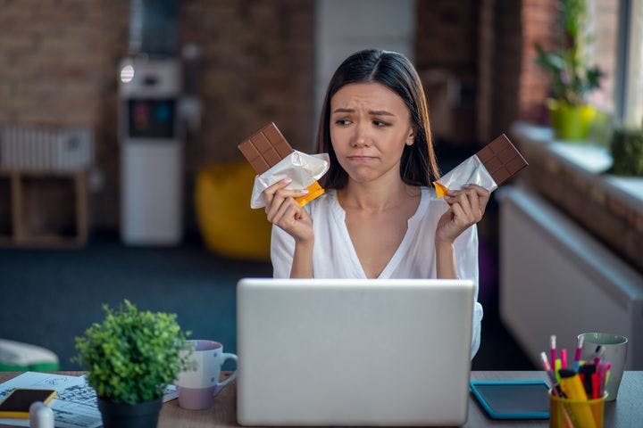 A woman sitting in front of her work desk while looking worriedly at the two chocolate bars in her hands.