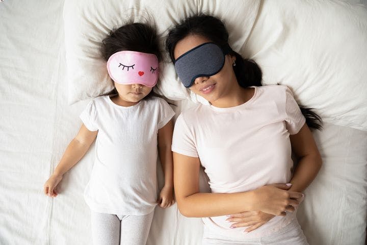 An overhead shot of a mother and her daughter sleeping side by side while wearing eye masks.