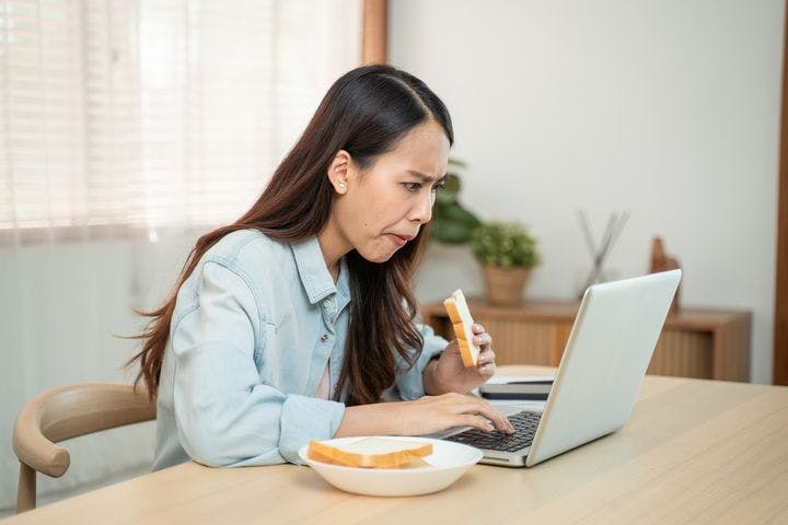 A woman staring hard at a laptop while eating a piece of bread.