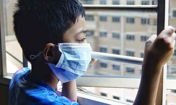 Boy wearing a blue-coloured medical mask as he looks out an open window
