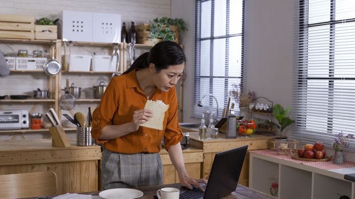 A woman standing while holding bread in one hand and looking at a laptop on a desk.