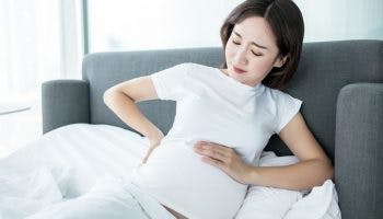 A pregnant woman sitting up in bed while holding her abdomen and lower back.