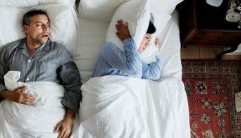 An man sleeping and snoring in bed with a woman lying sideways, covering her ears with a pillow.